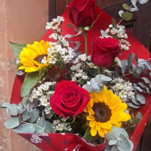 Mazzo di fiori con 3 rose rosse e 2 girasoli incorniciati da verde di stagione e confezionati con carta elegante e nastro.